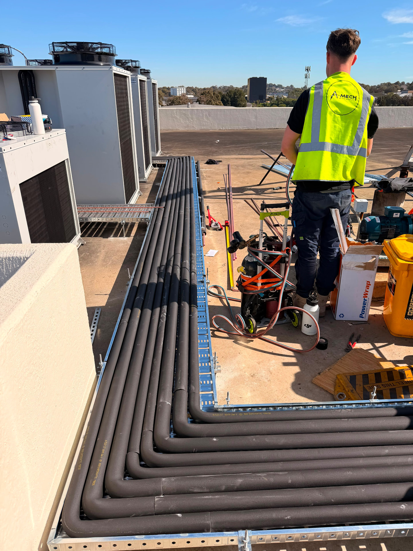 A-Mech tradesperson installing insulated refrigerant pipework on a rooftop beside condensing units